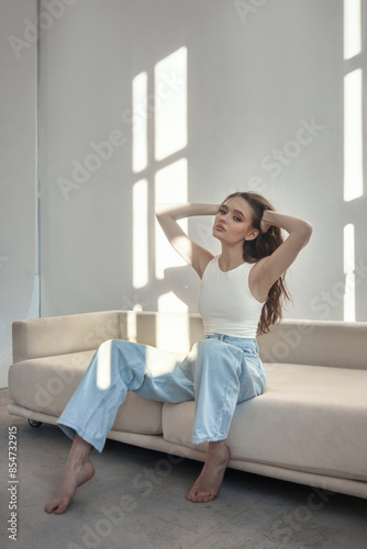Young fashion woman sits on a couch in a well-lit room. She is wearing blue jeans and a white tank top. The light shines through the window behind her Free Download Young fashion woman sits on a couch in a well-lit room. She is wearing blue jeans and a white tank top. The light shines through the window behind her - GFXLoad