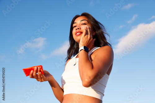  mixed race woman applying sunscreen to her face. It is a bright sunny summer day. She is smiling as she rubs in the sunscreen.