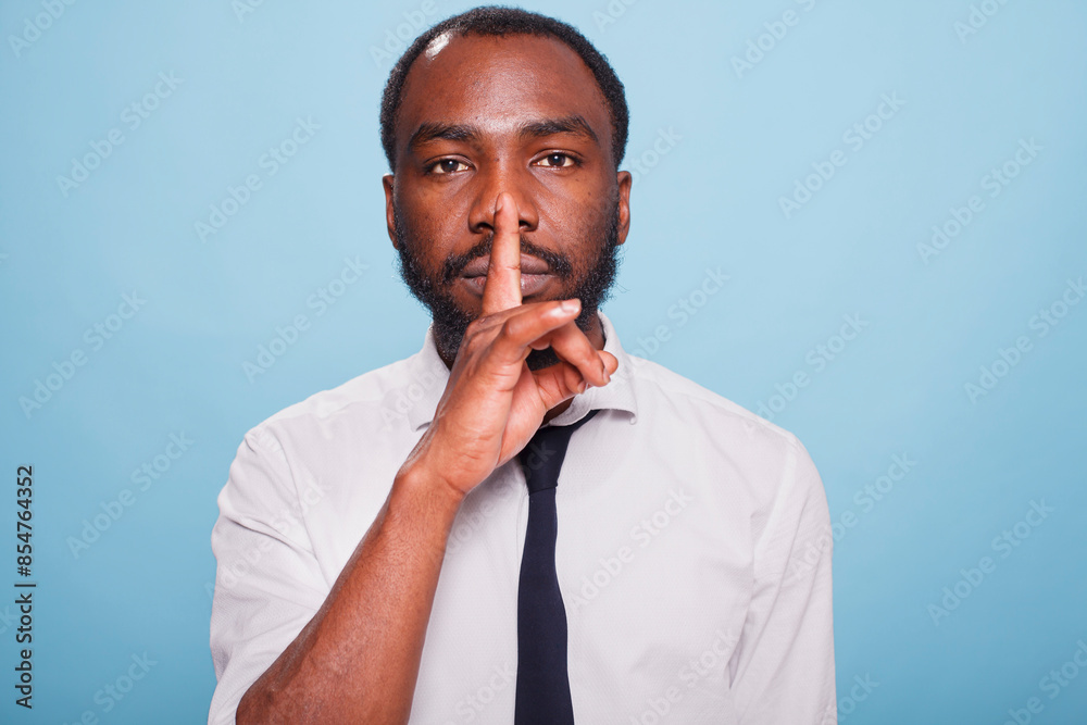 Portrait of african american male entrepreneur doing hush sign with index finger over lips on blue background. Calm professional teacher asking for silence by doing shush hand gesture. Portrait of african american male entrepreneur doing hush sign with index finger over lips on blue background. Calm professional teacher asking for silence by doing shush hand gesture.