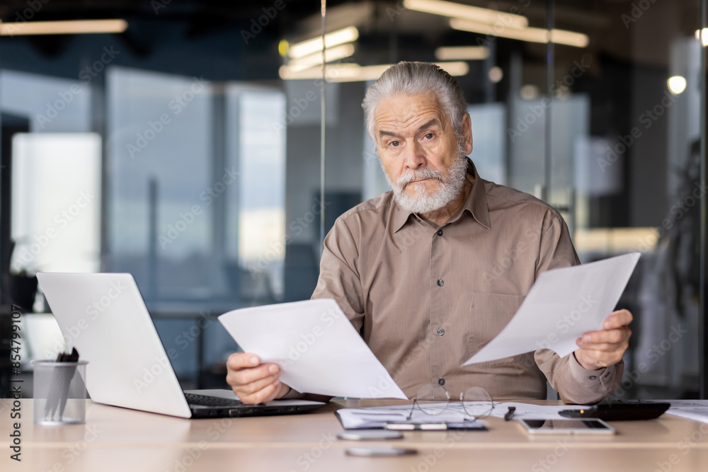 Senior businessman with gray beard reviewing documents in modern office setting, focused and serious. Working at a desk with laptop and papers. Senior businessman with gray beard reviewing documents in modern office setting, focused and serious. Working at a desk with laptop and papers.