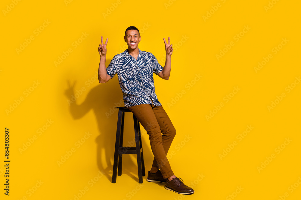 Fototapeta premium Full length photo of cheerful positive guy dressed print shirt sitting stool showing v-signs isolated yellow color background