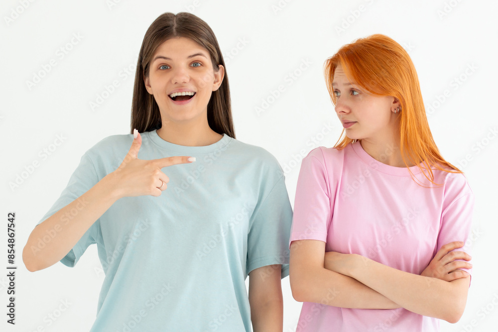 Portrait of two cheerful young women standing together, one girl point at the other, the other one is unhappy isolated over white background Portrait of two cheerful young women standing together, one girl point at the other, the other one is unhappy isolated over white background