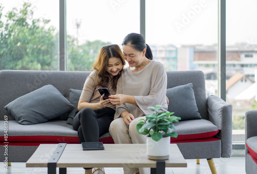 Two asian women sitting on a couch, smiling and looking at a mobile phone together.