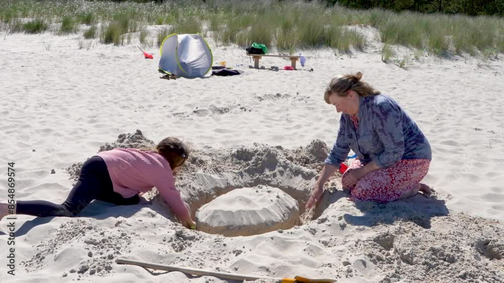 Grandmother and granddaughter play on the beach, building a large sand tower, digging sand on the beach with a shovel. Baltic Sea, Germany. Holidays with children, holidays Grandmother and granddaughter play on the beach, building a large sand tower, digging sand on the beach with a shovel. Baltic Sea, Germany. Holidays with children, holidays