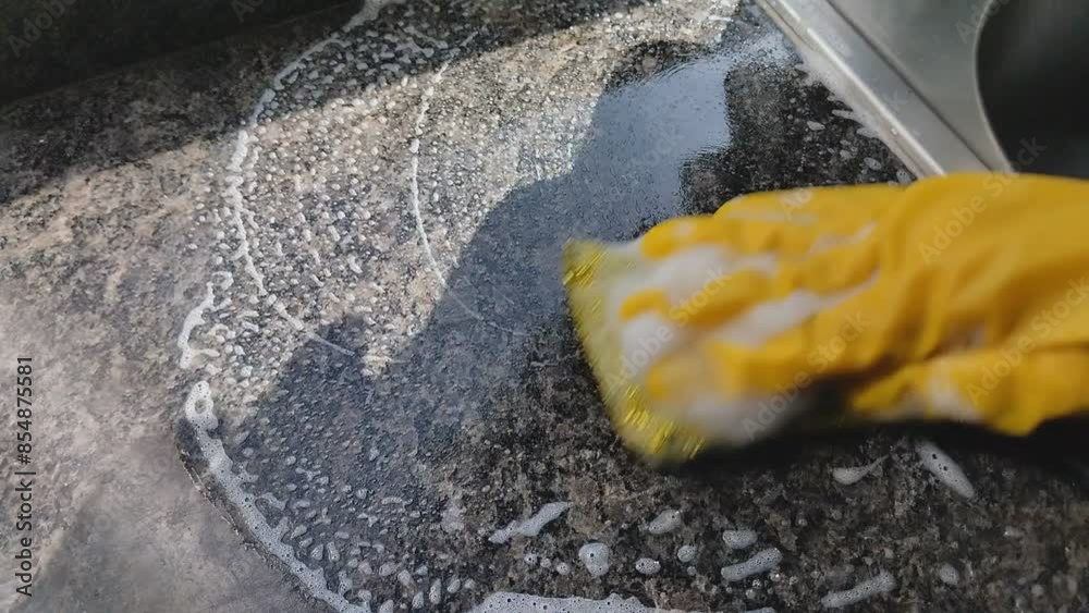Primer plano de una mano de mujer con un guante amarillo y una esponja con jabón limpiando la cocina de su casa.