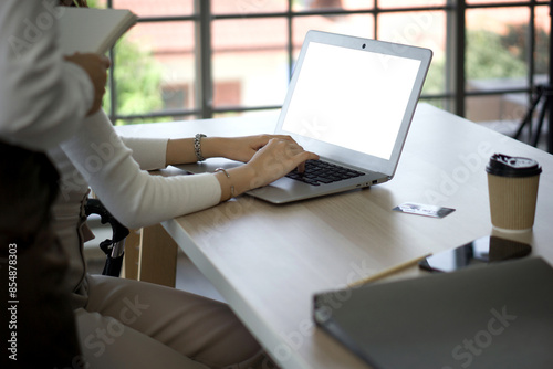 A person typing on white screen laptop computer in an office with coffee, credit card and file folder on the desk. There was a work supervisor standing behind her holding a notebook.