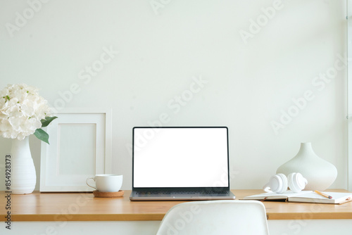 Front view laptop with blank white screen, books, coffee cup and picture frame on wooden table.