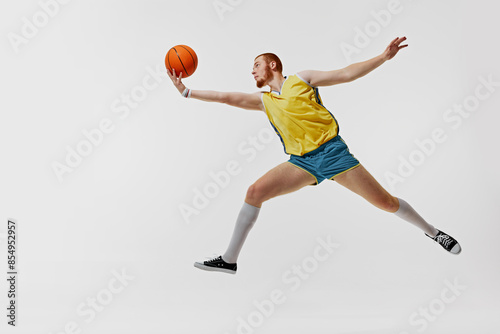 Dynamic image of young red haired man, basketball player in yellow jersey and blue shorts in motion with ball isolated on white background. Concept of sport, retro and vintage, active lifestyle, hobby