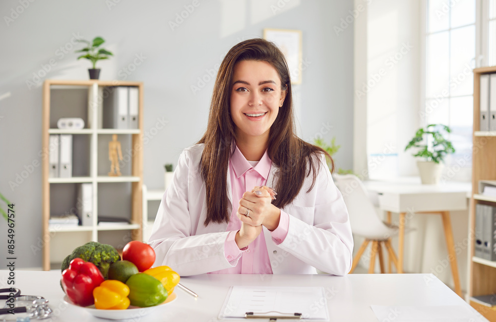 Portrait of a smiling doctor or nurse, a nutritionist, participating in video call, sitting at a table in a hospital and looking at the camera like a webcam, talking with patient or colleagues.
