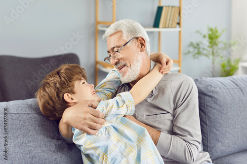 Portrait of cheerful senior grandfather with happy boy grandson sitting on sofa at home hugging and smiling. Elderly man having fun with child indoors enjoying time together. Family and love concept.