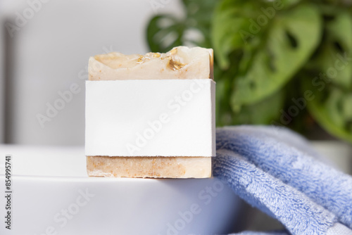 Soap bar with blank label on vessel sink near blue towels and plant close up, mockup photo