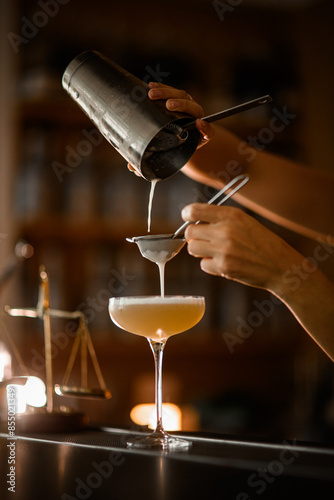 Female bartender finishes straining through two sieves a frothy champagne-colored cocktail into a stemmed glass photo