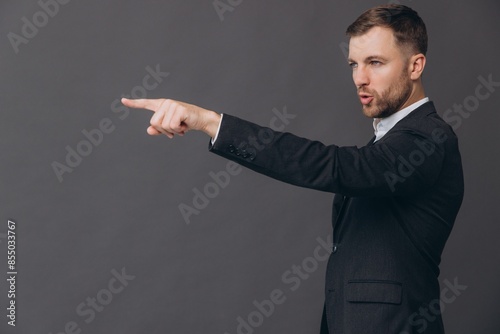 Caucasian millennial businessman isolated on gray studio background posing, nervous angry man in formal suit standing in profile pointing finger and saying you copy space