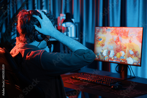 Host channel of stressful young gaming streamer getting to lose in fighting Moba at battle arena defeated multiplay team, wearing headphone on pc monitor with back side image at red neon room. Gusher. photo