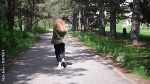Wallpaper Mural young woman jogs along shaded park path, dressed in green sports bra, olive green jacket, and black pants. She runs through a picturesque setting with trees and greenery on both sides, enjoying sunny Torontodigital.ca
