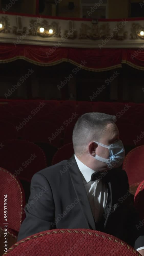 A lonely single man in a business suit and with a medical mask on his face is sitting in a chair in a theater hall. Vertical video. A lonely single man in a business suit and with a medical mask on his face is sitting in a chair in a theater hall. Vertical video.