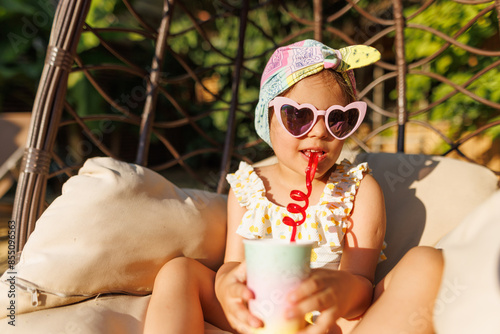 Funny cute girl drinks a cocktail on summer vacation. The child has fun on the beach. Cute baby in a colorful swimsuit and sunglasses is resting.