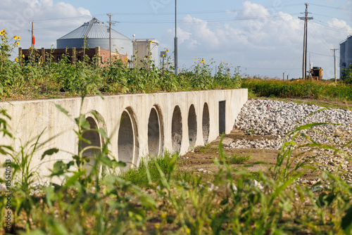 A row of concrete drainage ditches in a rural industrial area surrounded by weeds, sunflowers, and gravel with a silo in the background.