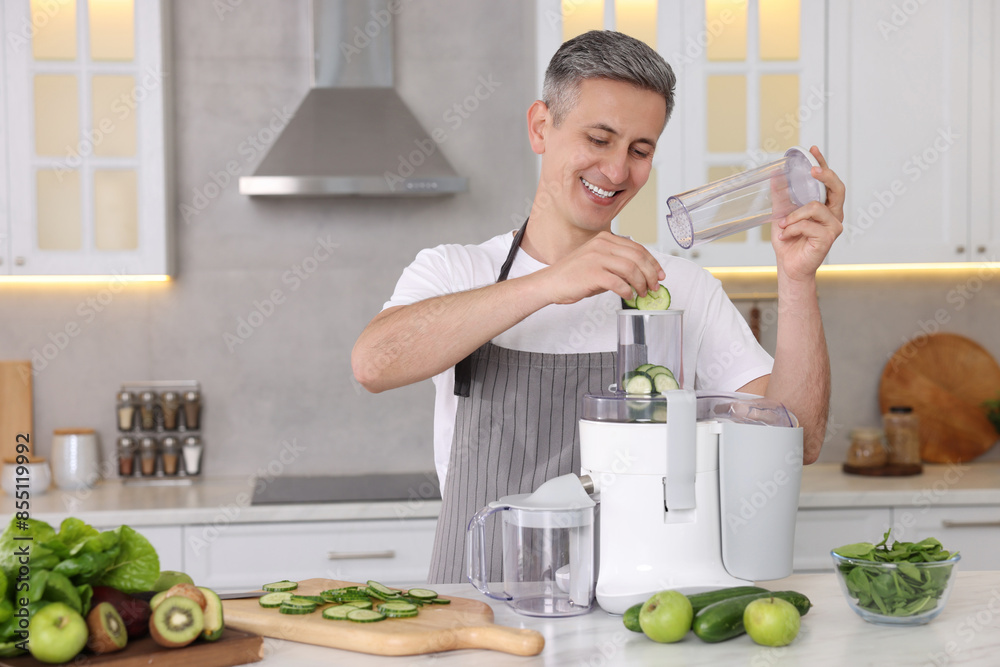 Smiling man putting fresh cucumber into juicer at white marble table in kitchen