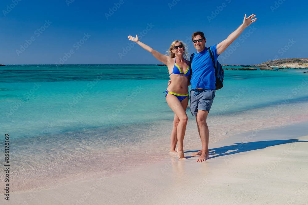 A man and a woman are smiling while posing for a photo at the beach. The background features the ocean, sky, sand, and joyous expressions. Pink sand. Pink shells. Elafonisi. Crete. Greece.