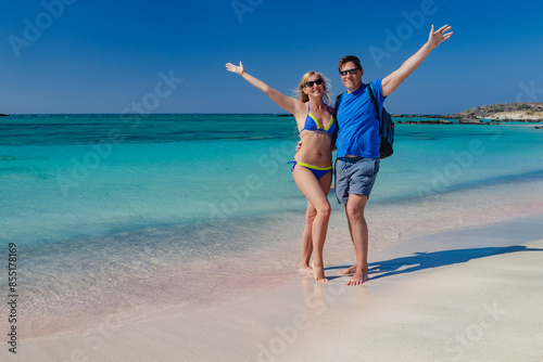 Obraz na płótnie A man and a woman are smiling while posing for a photo at the beach. The background features the ocean, sky, sand, and joyous expressions. Pink sand. Pink shells. Elafonisi. Crete. Greece. w salonie