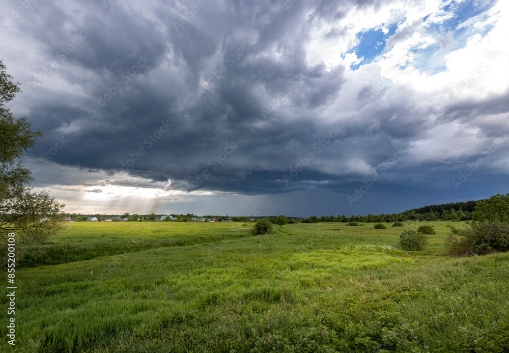 A dramatic view of storm clouds gathering over a lush green field. Sunlight pierces through the dark clouds, illuminating the landscape with an ethereal glow.