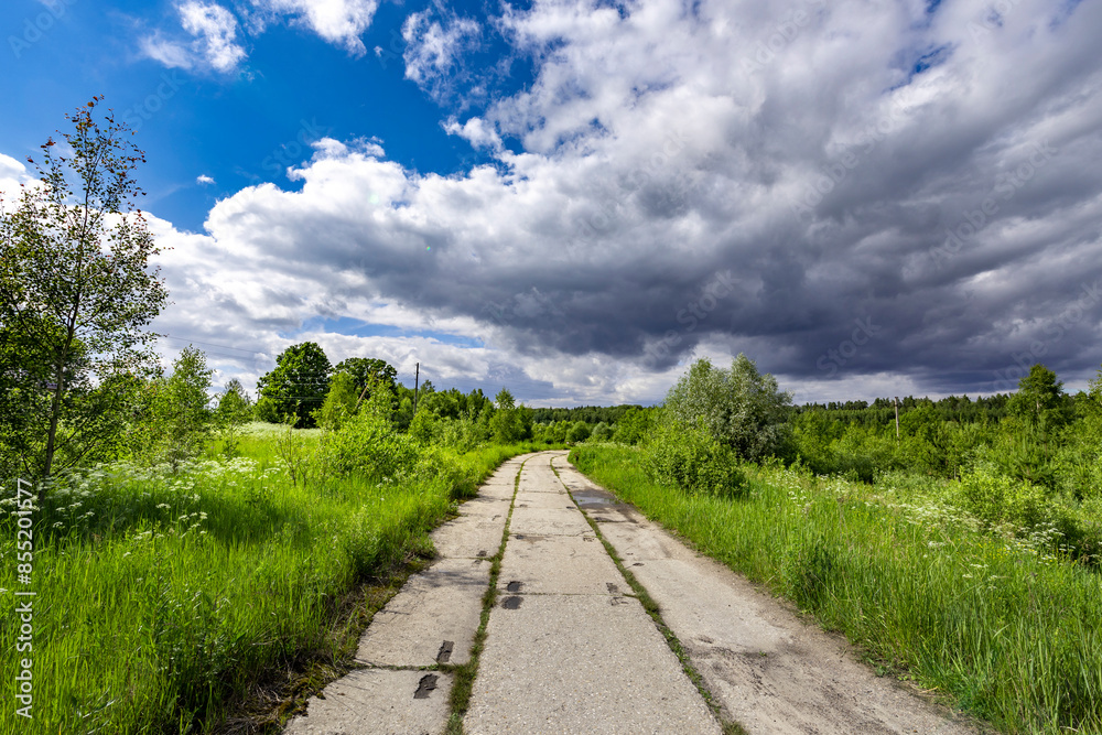A scenic road winds through lush green meadows and fields, disappearing into a distance where a dark, ominous storm cloud looms over the horizon.. A scenic road winds through lush green meadows and fields, disappearing into a distance where a dark, ominous storm cloud looms over the horizon..