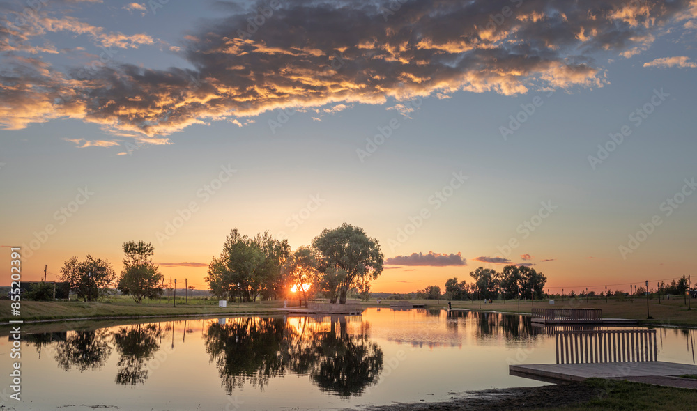 Obraz premium A serene sunset paints the sky with hues of orange and pink, reflecting off the tranquil waters of a park lake. Trees line the shore, creating a picturesque backdrop for a peaceful evening.