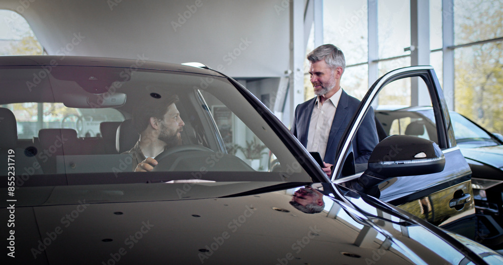 Caucasian buyer sitting inside new purchased automobile looking around in auto salon. Salesman manager with tablet is telling interested buyer about luxurious car. Caucasian buyer sitting inside new purchased automobile looking around in auto salon. Salesman manager with tablet is telling interested buyer about luxurious car.