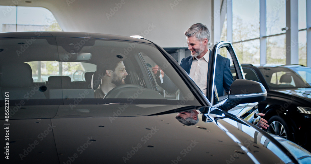Bearded caucasian male manager giving keys from new car to happy male client. Young caucasian man sitting in auto and feeling happiness from expensive purchase. Bearded caucasian male manager giving keys from new car to happy male client. Young caucasian man sitting in auto and feeling happiness from expensive purchase.