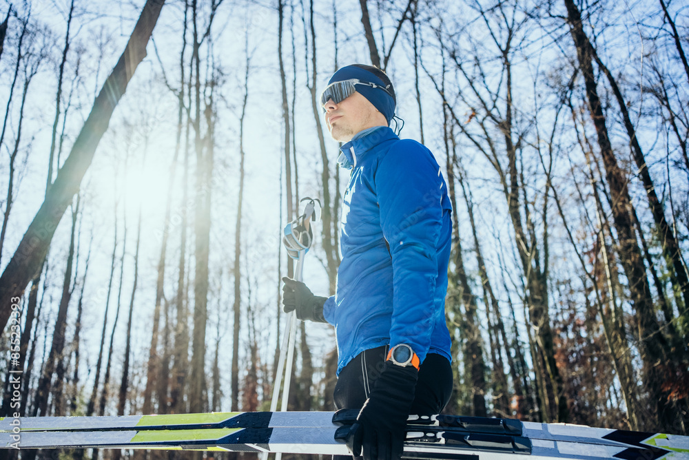 Cross Country Skier in Blue Jacket Enjoying a Sunny Winter Day in the Forest. Man dressed in blue winter sportswear is skiing through winter forest. He is wearing sunglasses and a watch, holding ski Cross Country Skier in Blue Jacket Enjoying a Sunny Winter Day in the Forest. Man dressed in blue winter sportswear is skiing through winter forest. He is wearing sunglasses and a watch, holding ski