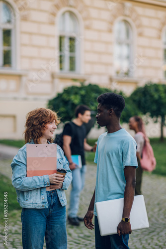 Diverse exchange student with laptop chatting with classmate at campus