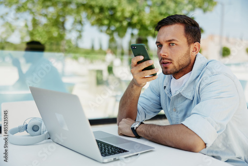 Man in casual clothing using a smartphone for voice command while working on a laptop outdoors. photo