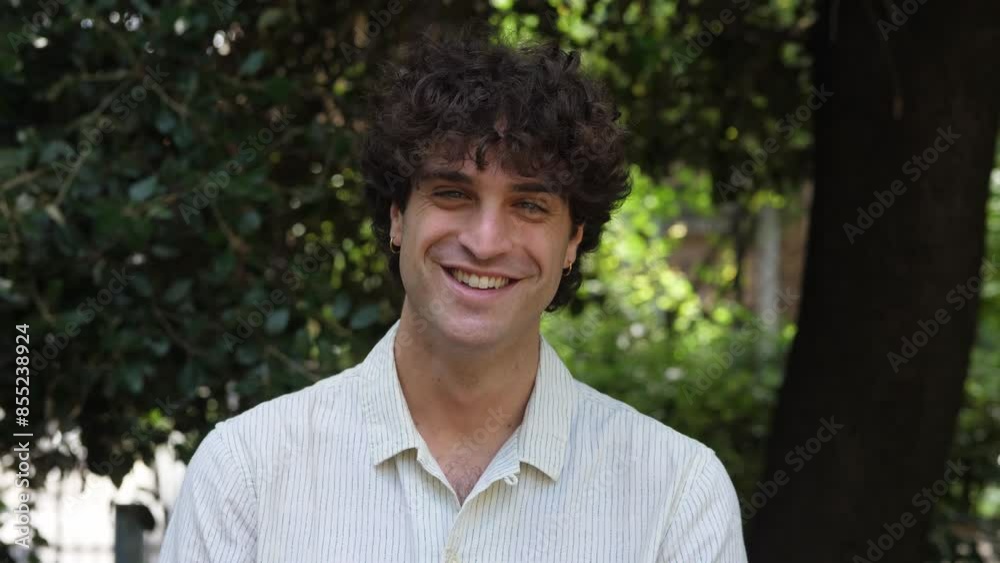 A young man with curly short hair and a light shirt smiles warmly at the camera while standing in a park, capturing a moment of joy and friendliness A young man with curly short hair and a light shirt smiles warmly at the camera while standing in a park, capturing a moment of joy and friendliness