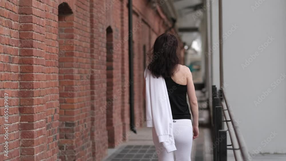 A woman in a white tracksuit walks through a narrow alley with red brick walls, heading towards her training session. The industrial and urban environment underlines her commitment to maintaining an A woman in a white tracksuit walks through a narrow alley with red brick walls, heading towards her training session. The industrial and urban environment underlines her commitment to maintaining an