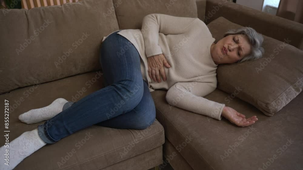 Elderly woman experiencing abdominal pain while lying on a couch. She is visibly uncomfortable and holding her stomach. Elderly woman experiencing abdominal pain while lying on a couch. She is visibly uncomfortable and holding her stomach.