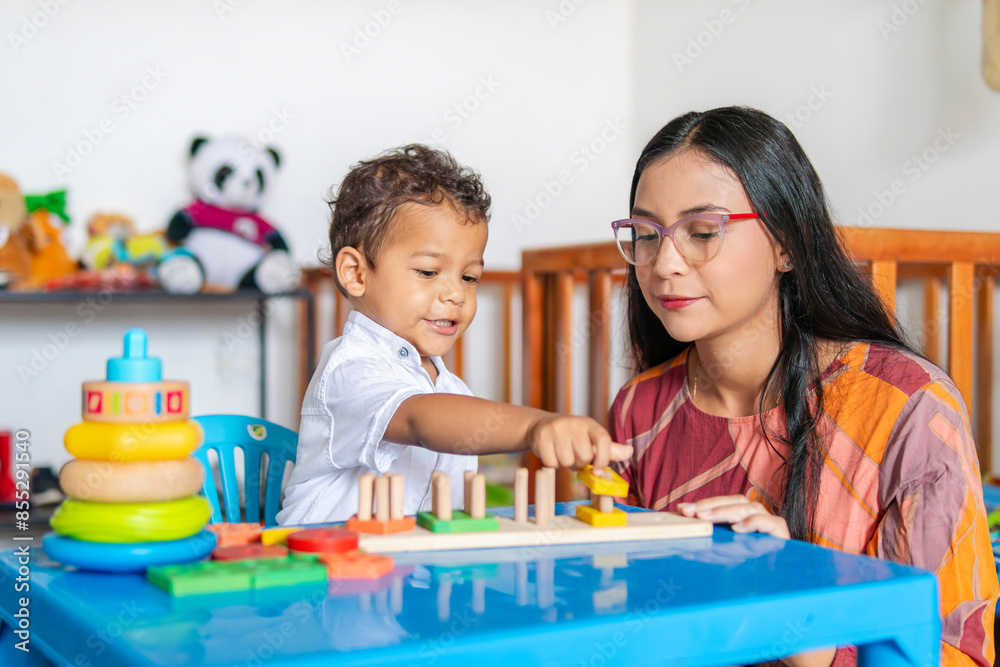Young Latina mother in her 20s, single, playing with her 2-year-old son in the nursery, where the crib and various toys are visible in the background Young Latina mother in her 20s, single, playing with her 2-year-old son in the nursery, where the crib and various toys are visible in the background
