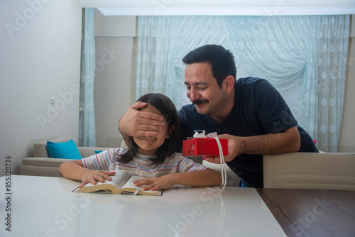 The father who closes his daughter's eyes while she is reading a book and gives her a red gift box