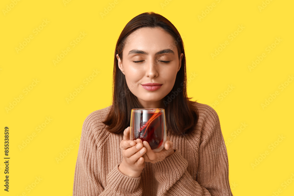 Beautiful young woman with glass cup of hot mulled wine on yellow background
