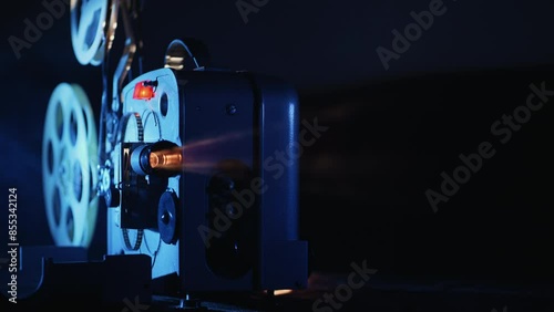 A vintage movie projector in operation. Blue light illuminates the film reels, creating a cinematic atmosphere. The background is dark, highlighting the projector photo