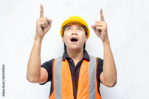 asian man consrtruction worker wearing yellow safety helmet and orange vest smiling and pointing finger to copy space with happy and surprised expression. photo