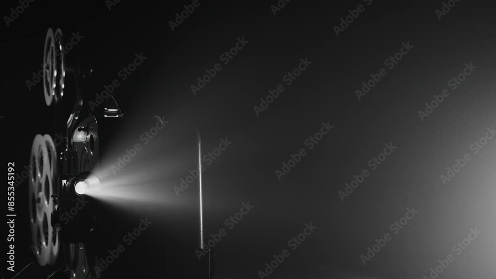 A black-and-white vintage movie projector working in the dark. A bright beam of light shines through the lens, illuminating the film reels. The atmosphere evokes classic film screenings
