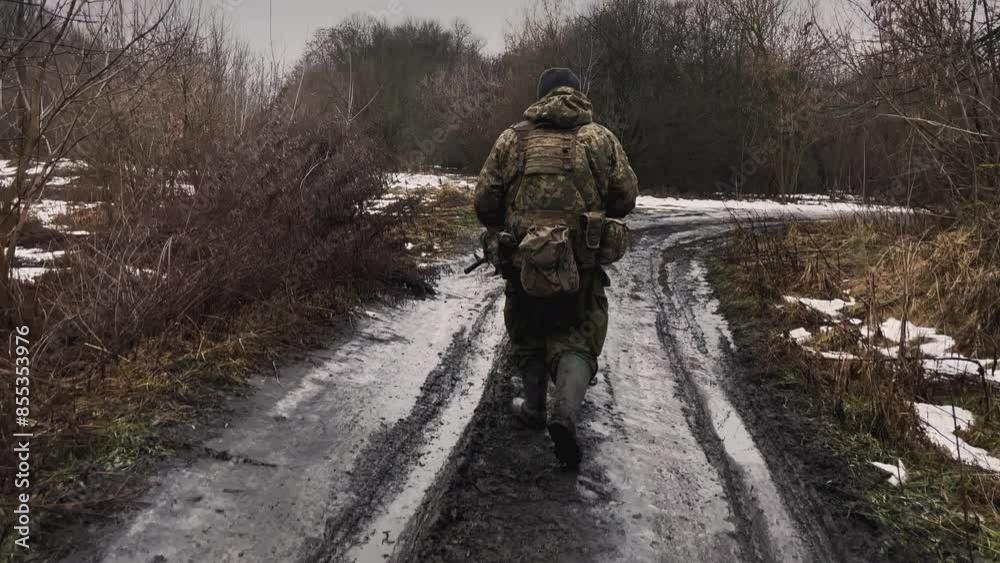 A Ukrainian soldier walks down a muddy road in a forest area. He is dressed in military uniform and carries a backpack and gear. There are remnants of melting snow on the ground A Ukrainian soldier walks down a muddy road in a forest area. He is dressed in military uniform and carries a backpack and gear. There are remnants of melting snow on the ground