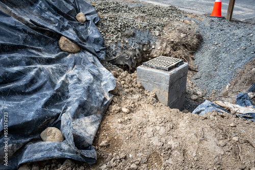 New storm water sewar installation in progress, new cement pore with rectangular iron grate on top, hole dig in dirt with partial gravel, rock, and dirt backfill
 photo