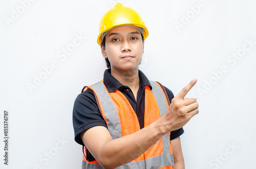 asian man consrtruction worker wearing yellow safety helmet and orange vest smiling and pointing finger to copy space with happy and surprised expression.