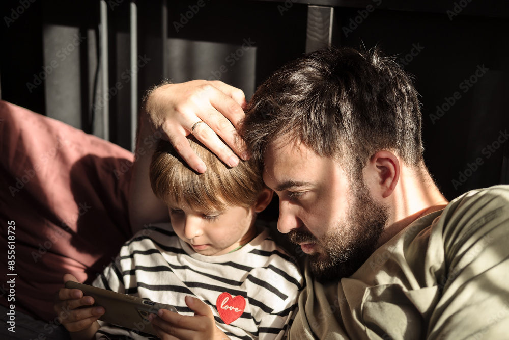 Obraz premium Son and dad are having a great time together. They are looking at the smartphone together and hugging. Concept of a happy childhood and parenthood.