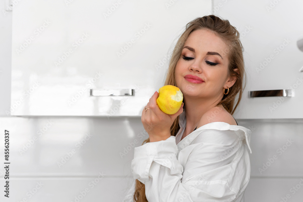 Beautiful young woman in white shirt eating yellow apple in kitchen. Healthy eating. Beautiful young woman in white shirt eating yellow apple in kitchen. Healthy eating.