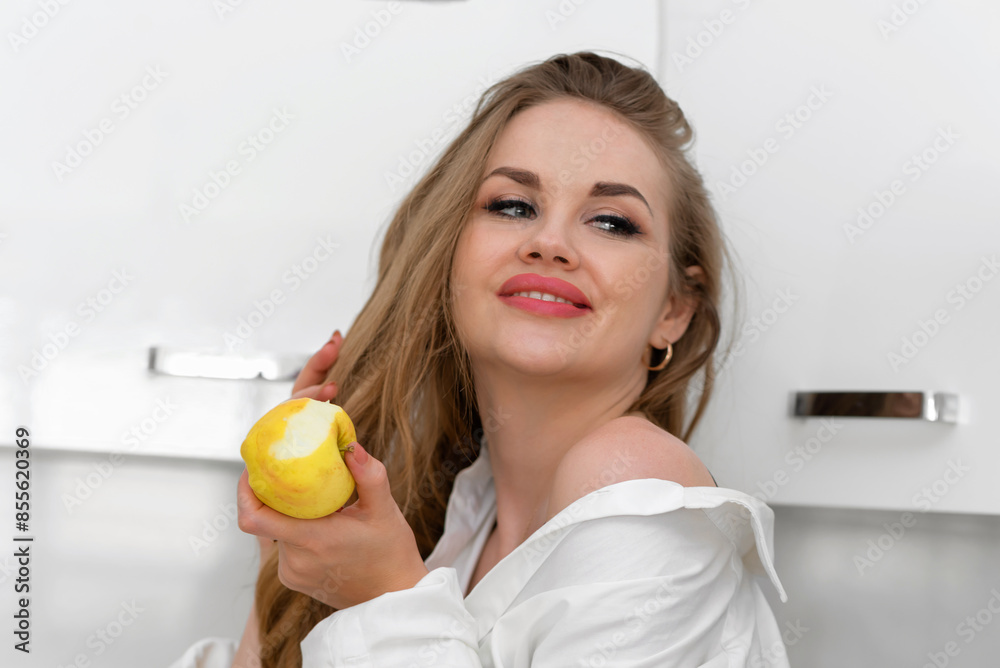 Beautiful young woman in white shirt eating yellow apple in kitchen. Healthy eating. Beautiful young woman in white shirt eating yellow apple in kitchen. Healthy eating.