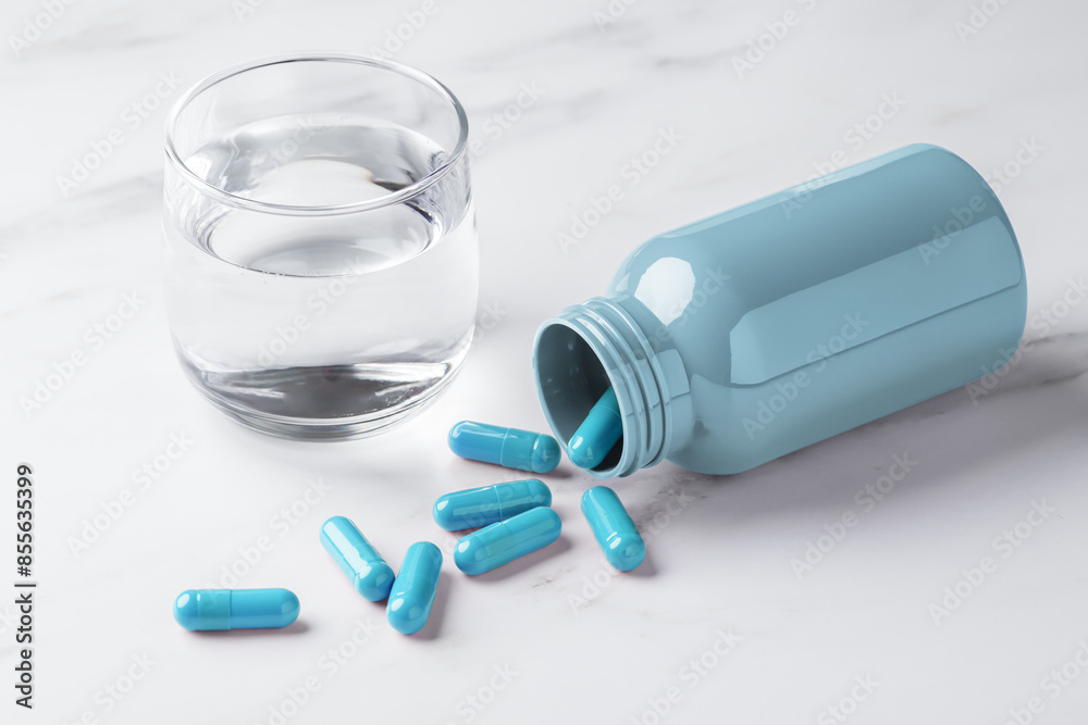 Blue pill capsules spilling out of bottle and glass of water on white table background