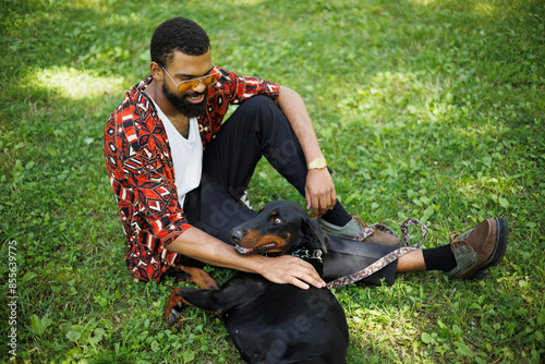 Relaxed african american pet owner sitting near dog on grass in summer park © Dmytro Hai